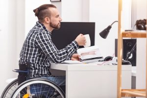 disabled man in a wheelchair drinking coffee and holding a book at his desk
