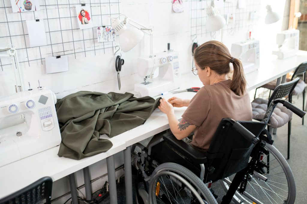 Young disable female in casualwear sitting in wheelchair by workplace inside contemporary workshop