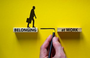 Belonging at work symbol. Wooden blocks with words 'Belonging at work' on beautiful yellow background. Businessman hand. Businessman icon. Business, belonging at work concept.