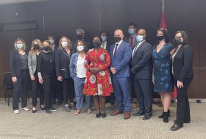 Day on the Hill, group photo. Fourteen people of mixed genders, ages, and races together in colorful business attire. They stand in a conference room with tan floor and brown wood-grain walls, and a Canadian flag is behind them. They all wear medical masks.