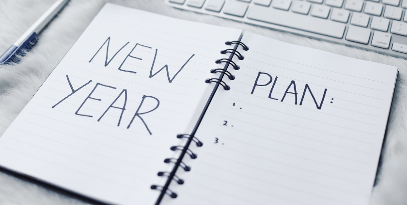 An open notepad rests on a desk beside a pen and keyboard. The open pages read “New Year Plan,” with open space to fill in.