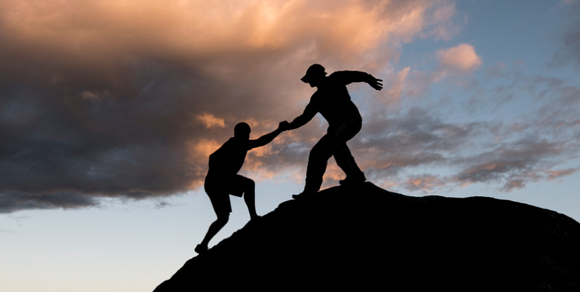 The silhouette of two hikers atop a rocky hill at sunset. One extends a helping hand to other, reaching the top together.