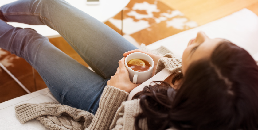 A woman leans back into a comfortable sofa. She holds a warm cup of tea with a slice of lemon.