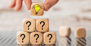 Photo: Wooden blocks printed with question marks are being stacked into a pyramid. A hand places the topmost block printed with a lightbulb.
