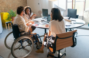 Photo: In an office space, three adults - one sitting in a wheelchair - have an energetic conversation at a table covered in papers and laptops.