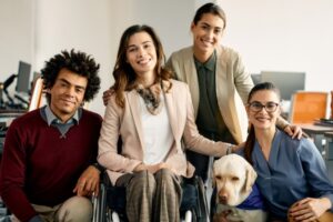 A photo of a group of young professionals huddled together. Dressed for business, they are diverse in ethnicity and some display accessibility aids such as a wheelchair and service dog.